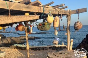 Das Bild zeigt eine rustikale Holzbude am Strand mit allerlei Strand-Utensilien mit Blick aufs Meer als Bildmetapher für eine Reflexion zur Sommerzeit (Photocredit: Petra Welz, Geld & Rosen, Düsseldorf)
