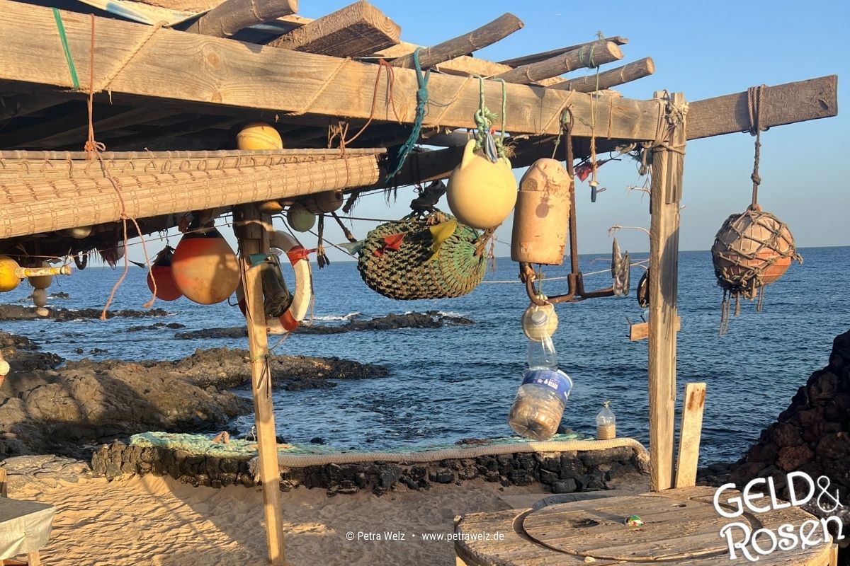 Das Bild zeigt eine rustikale Holzbude am Strand mit allerlei Strand-Utensilien mit Blick aufs Meer als Bildmetapher für eine Reflexion zur Sommerzeit (Photocredit: Petra Welz, Geld & Rosen, Düsseldorf)