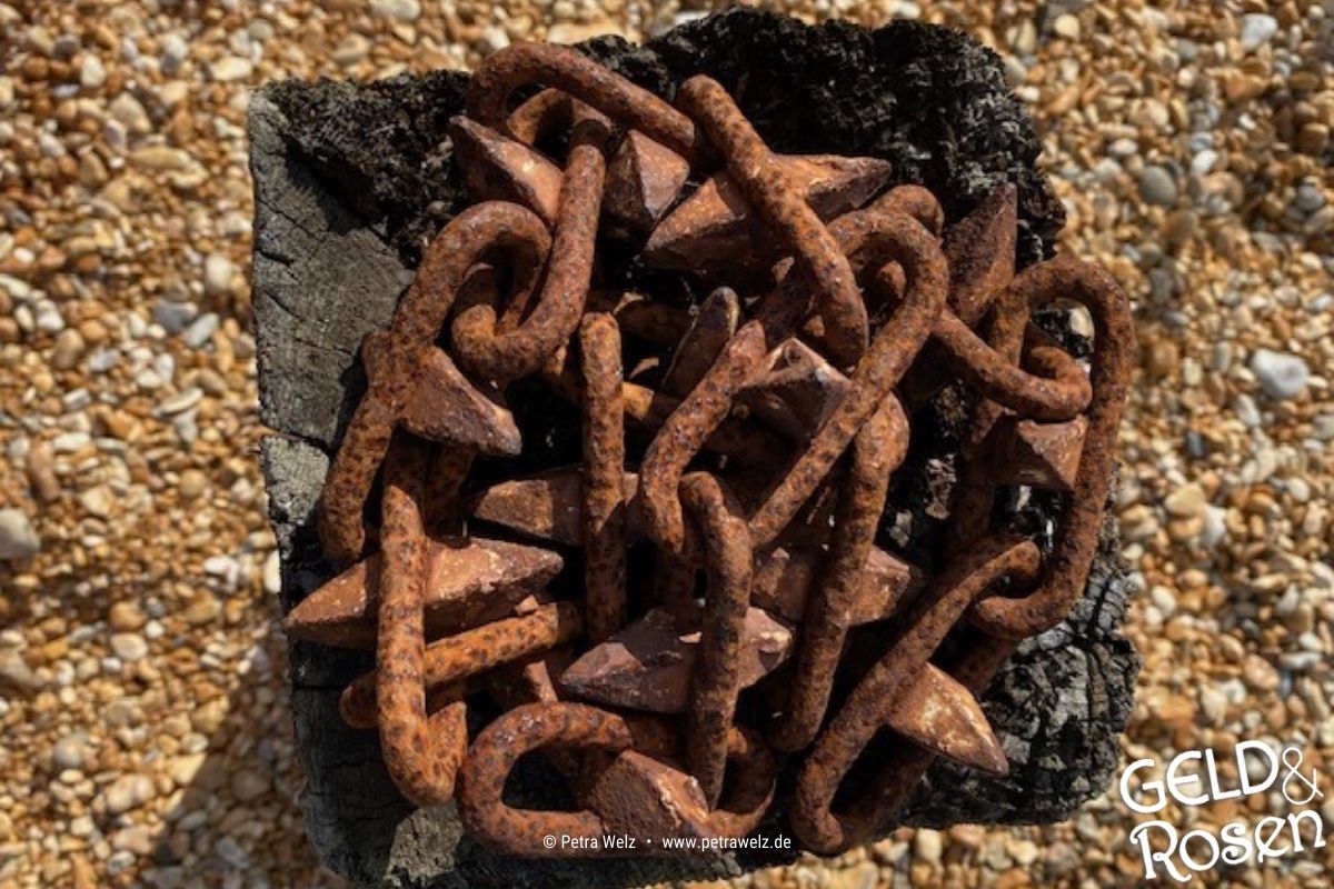 Das Bild zeigt eine verknotete Eisenkette, die in einer eckigen verkohlten Holzschale liegt – Analogie zur Wirtschaftlichkeit im eigenen Unternehmen (Photocredit: Petra Welz, Geld & Rosen, Düsseldorf)