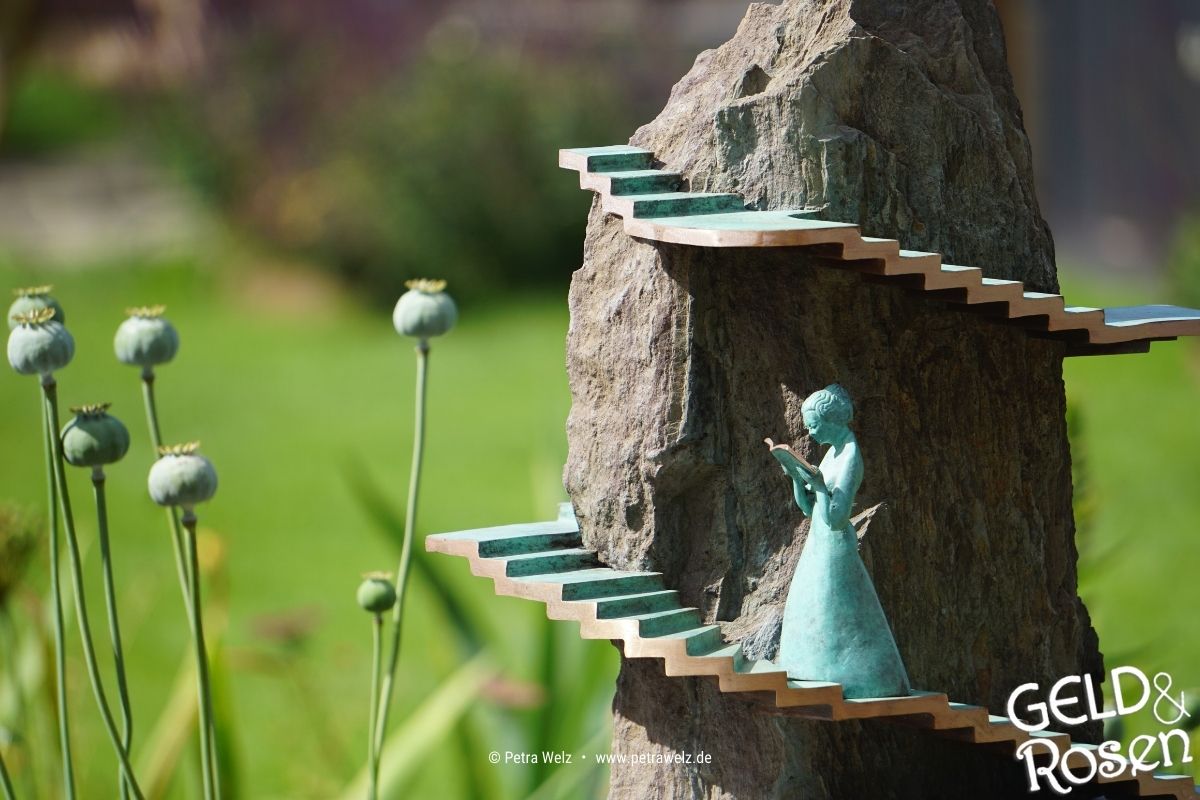 Das Bild zeigt einen Baumstein, um den eine kleine Treppe führt, auf der eine kleine lesende Figur platziert ist, die Selbstführung manifestiert (Photocredit: Petra Welz, Geld & Rosen, Düsseldorf).